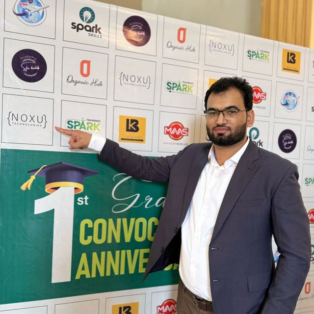 Muhammad Asif at a student convocation, pointing to the NOXU Technologies logo on the sponsor wall